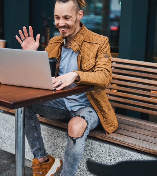 Happy young man with dreadlocks is sitting at table outdoors with cup of coffee and using camera on laptop for saying hello to friend